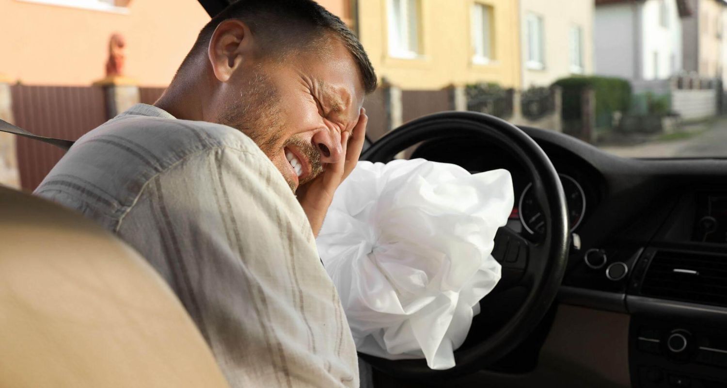 Man in a car grimacing in pain, head resting on his hand with a white tissue near the steering wheel.
