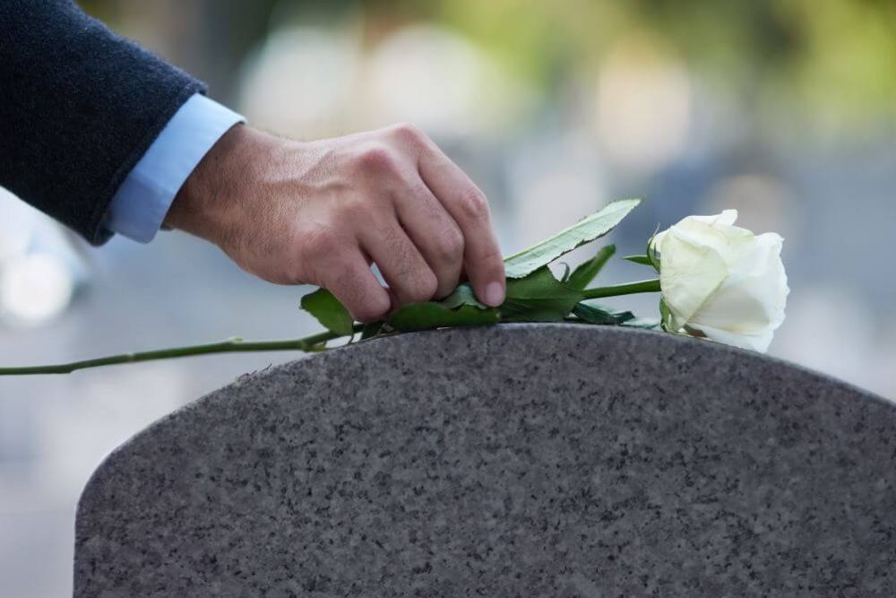 A person's hand placing a white rose on a granite surface, likely symbolizing remembrance or mourning, in an outdoor setting.