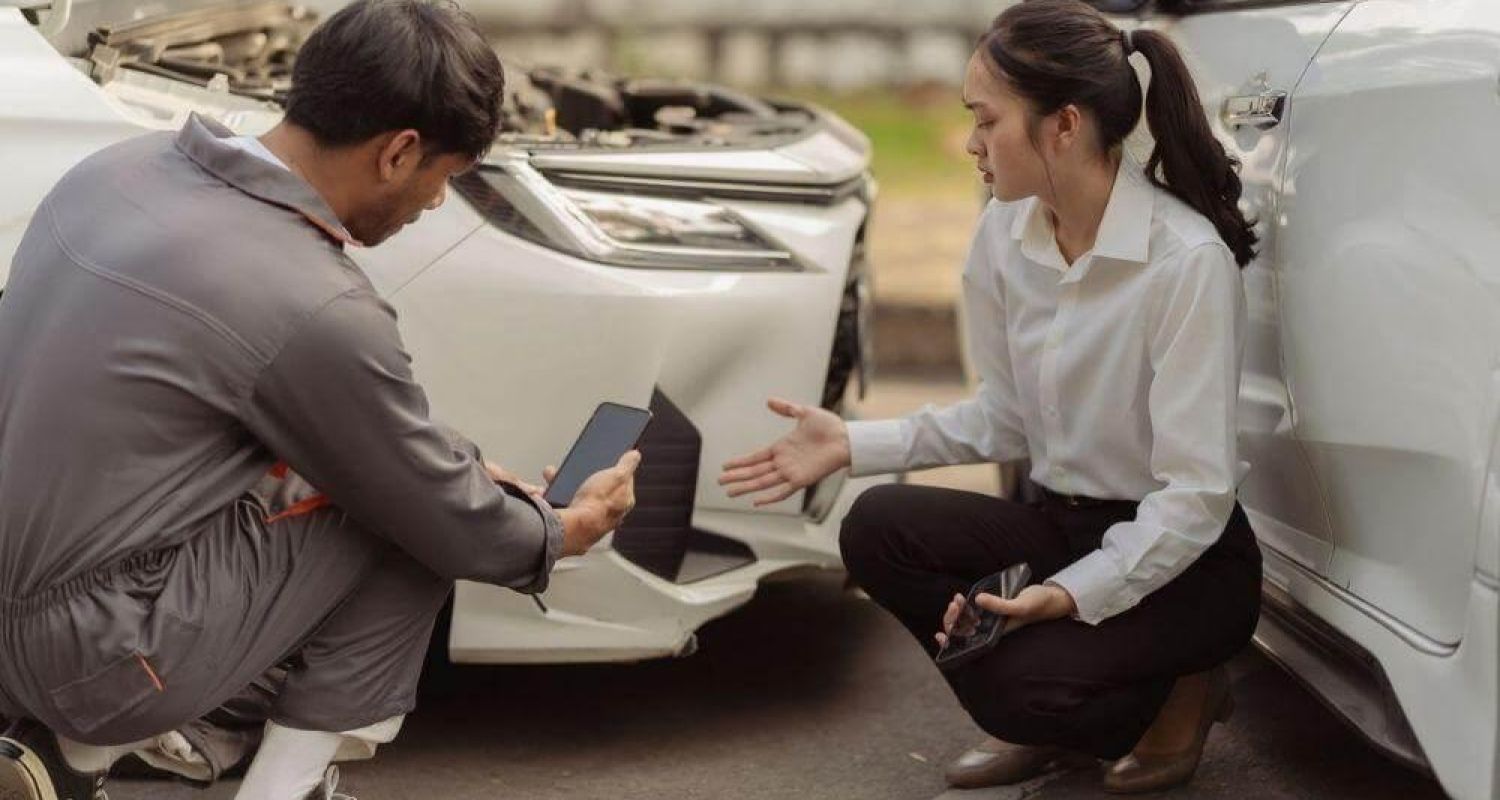 Two people kneel beside a white car with its hood open, discussing damage or repairs. One person holds a phone/tablet while the other gestures toward the car.