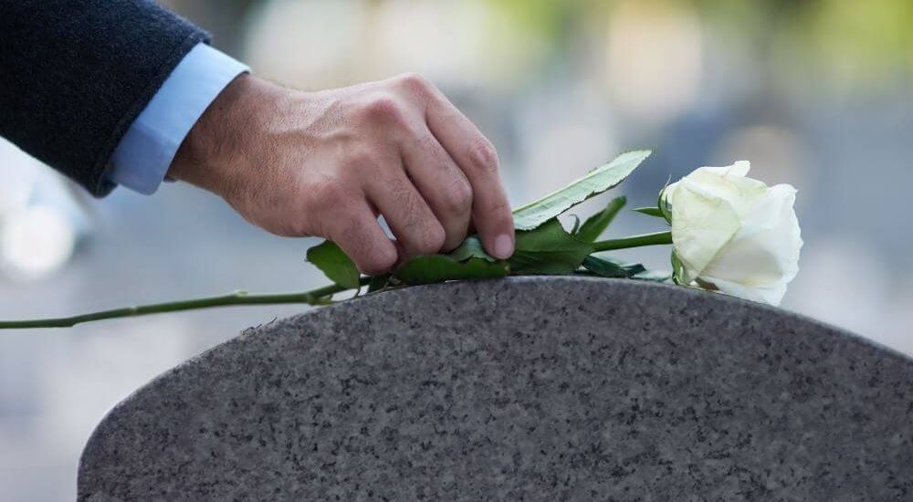 A person's hand placing a white rose on a granite surface, likely symbolizing remembrance or mourning, in an outdoor setting.