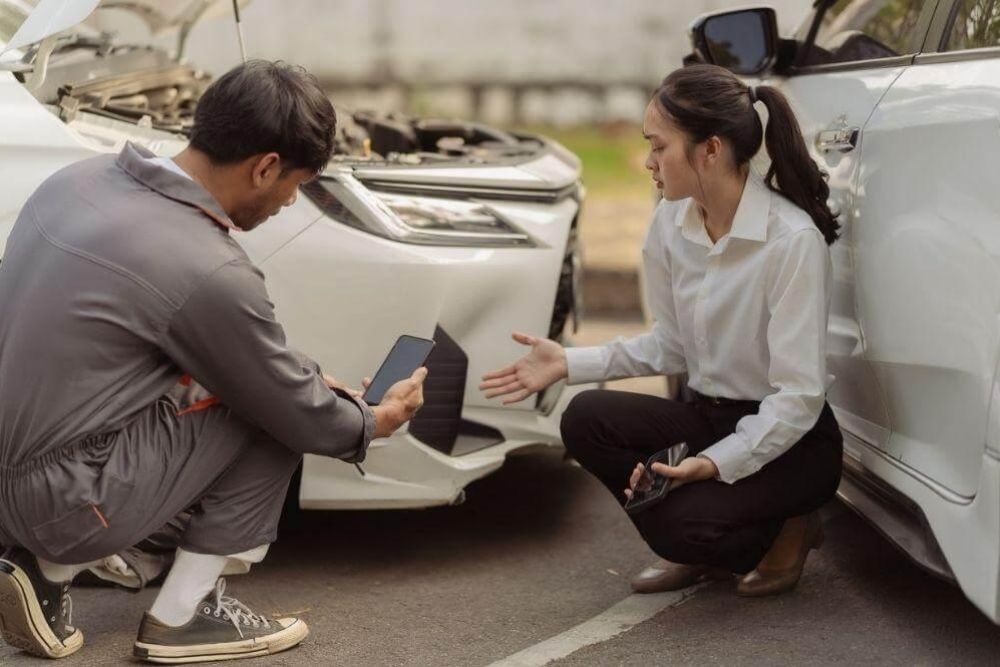 Two people kneel beside a white car with its hood open, discussing damage or repairs. One person holds a phone/tablet while the other gestures toward the car.